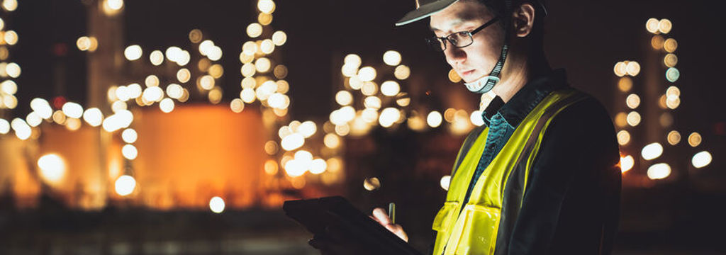 Worker in hard hat holding tablet in front of industrial plant, in the dark